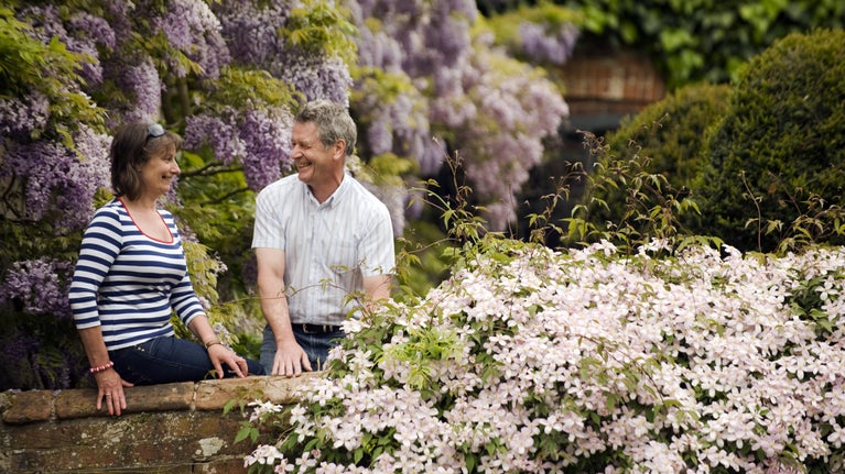 A man and a woman in the courtyard garden at Baddesley Clinton, Warwickshire. Behind them the wisteria is in full bloom, and a clematis is flowering